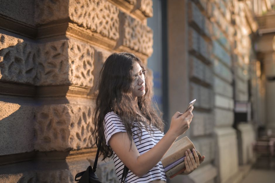 Casual portrait of a young woman leaning on a wall, using her smartphone and holding books outdoors.