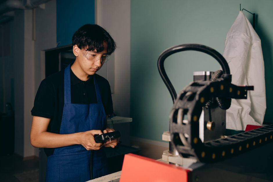 Asian man in a workshop setting operating a machine while wearing goggles and apron.