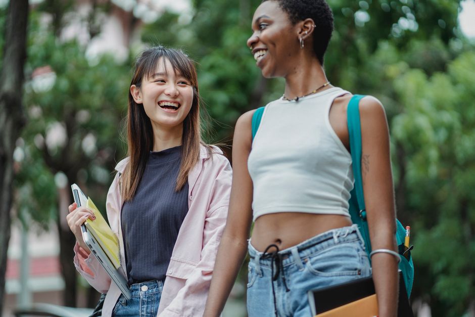Delighted multiracial female students in casual outfits carrying backpack and copybooks while strolling in lush park and discussing plans