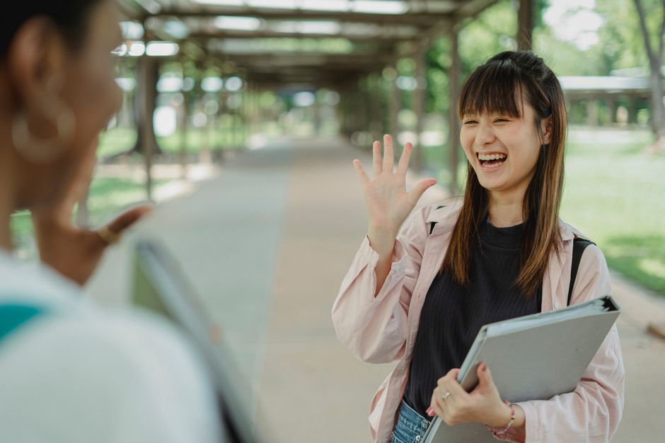 Young positive ethnic female student with folder greeting crop unrecognizable best friend on walkway while looking at each other