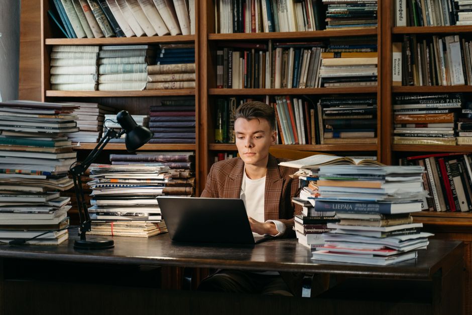 A young man intensively studying at a library surrounded by books, using a laptop.